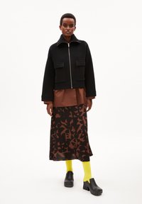 Chaqueta negra corta con cremallera y dos bolsillos frontales, sobre una camisa marrón y una falda larga con estampado. Calcetines amarillos brillantes y zuecos negros.