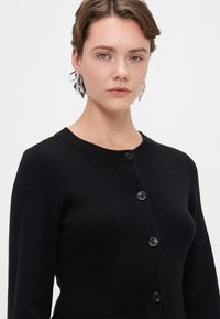 Μαύρο πλεκτό κασκόλ με στρογγυλή λαιμόκοψη, πέντε κουμπιά και διακριτική υφή. Ο σχεδιασμός είναι εφαρμοστός με τσέπες μπροστά.