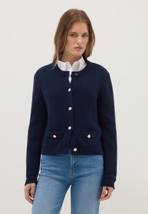 Woman wearing navy blue buttoned cardigan with two front pockets over white collared shirt and light blue jeans, standing against plain background.