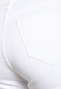 Los jeans de mezclilla blanca presentan un prominente bolsillo trasero con un diseño de costura distintivo. La textura parece suave y bien tejida.