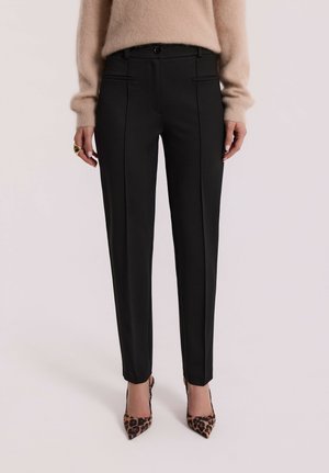 Kobieta w czarnych dopasowanych spodniach, beżowym puszystym swetrze, z butami na obcasie w panterkę oraz z złotym pierścionkiem na lewej ręce.