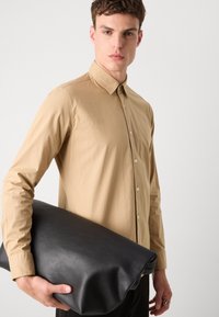 Light brown button-up shirt made of smooth fabric, held with one hand. The other hand supports a large, textured black leather bag.