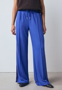 Kék, széles szárú nadrág, sima anyagból, gumis derékrésszel és húzózsinórral, kontrasztos oldalsávokkal.