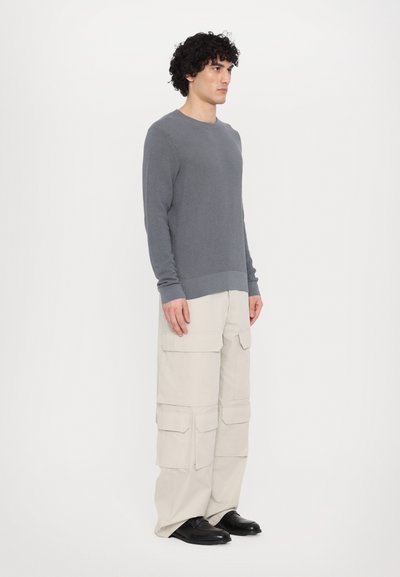 Grauer, langärmeliger Strickpullover mit gerippter Textur, kombiniert mit cremefarbenen Cargo-Hosen, die mehrere aufgesetzte Taschen haben. Schwarze Schuhe runden das Outfit ab.