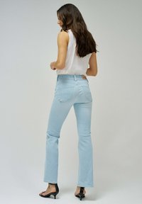 Jeans de denim de tiro alto, acampanados y de color azul claro, con cinco bolsillos y una etiqueta de cuero en la parte posterior de la cinturilla. Modelo con una camiseta sin mangas blanca.