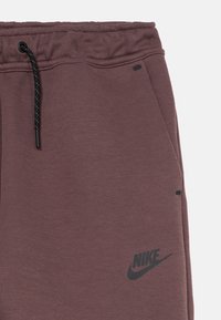 Tmavofialové tepláky so šnúrkou v páse, vybavené bočnými vreckami a čiernym logom Nike na ľavej strane. Hladká textúra.