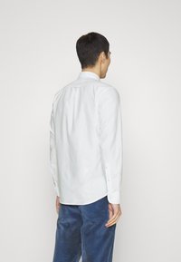 Man som bär en vit skjorta med lång ärm och blå kordbyxor, står vänd bort från kameran mot en enkel ljus bakgrund.