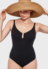 Maillot de bain une pièce noir à côtes avec un décolleté en V et un petit trou clé à l'avant. Le modèle porte un chapeau de paille à larges bords.
