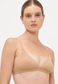 Vrouw met lichte huid en haar in een lage knot, gekleed in een naadloze driehoekige bh in huidskleur, staat rechtop tegen een effen lichte achtergrond.