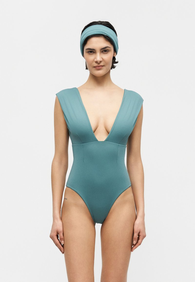 Γυναίκα που φοράει πετρόλ ολόσωμο μαγιό με βαθύ V-λαιμό χωρίς μανίκια και ταιριαστή κορδέλα, στέκεται μπροστά σε απλό λευκό φόντο.