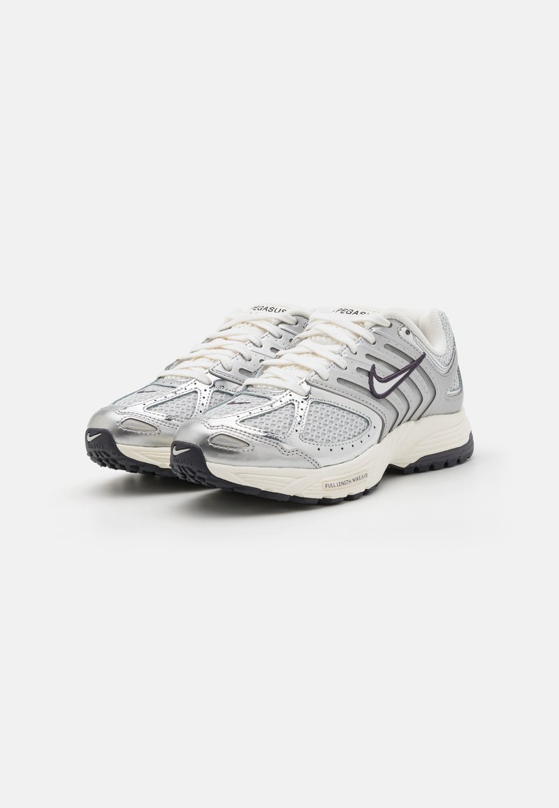 Zapatillas atléticas plateadas y blancas con parte superior de malla, acentos texturizados, lengüeta acolchada y suela de goma negra. Presenta cordones y detalle de logo.