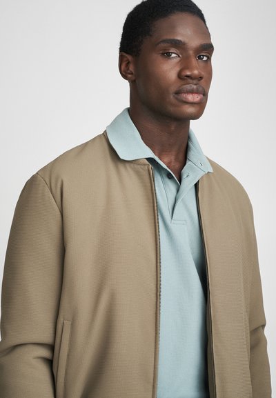 Καφέ υφασματική bomber ζακέτα πάνω από ένα ανοιχτό μπλε πόλο πουκάμισο με γιακά. Η ζακέτα έχει φερμουάρ και πλαϊνές τσέπες.