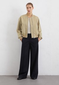 Veste bomber beige avec une fermeture éclair à l'avant et des poignets côtelés, associée à un pantalon noir à jambes larges. Tissu lisse avec un design décontracté.