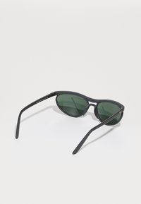 Óculos de sol pretos com lentes verdes. Formato oval, armação fina e hastes curvas. Acabamento texturizado na armação. Design minimalista.