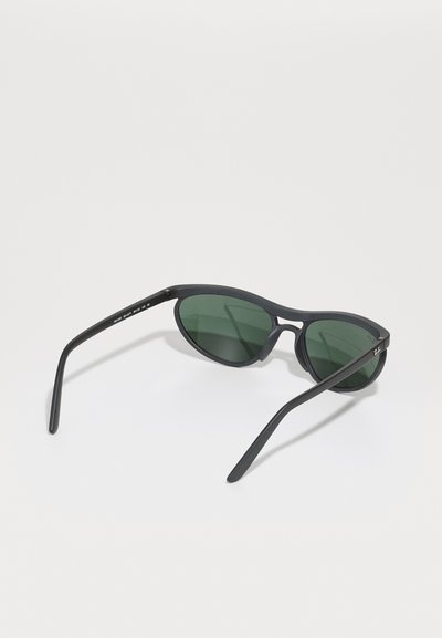 Lunettes de soleil noires avec des verres verts. Forme ovale, monture fine et branches courbées. Fini texturé sur la monture. Design minimaliste.
