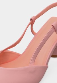 Κοντινό πλάνο ροζ παπουτσιού με τακούνι και λεπτό λουράκι στο πίσω μέρος, με μυτερή μύτη, σε απλό φόντο.