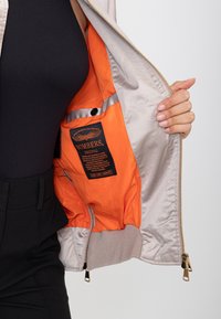 Main tenant une veste beige ouverte révélant une doublure orange vif et une étiquette intérieure avec le texte "BOMBERS ORIGINAL" sur tissu noir.