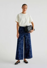 Pantalons larges en denim bleu foncé à motif dans un tissu texturé, associés à un T-shirt uni de couleur claire et à un sac à main noir. Chaussures plates noires.