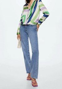 Blusa di seta multicolore con motivi geometrici verdi, viola e blu, abbinata a jeans di denim blu chiaro e sandali viola con cinturini.