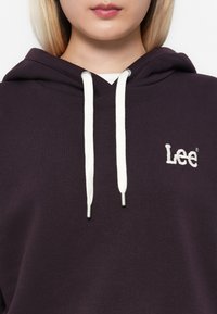 Haut à capuche violet avec un logo brodé blanc "Lee" et des cordons de couleur crème avec des embouts en métal. Tissu doux en mélange de coton.