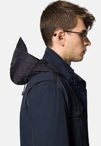 Granatowa kurtka z kapturem o eleganckim fasonie, wykonana z lekkiego materiału, z zatrzaskami i teksturowanym kapturem. Widok z boku.