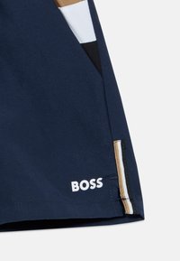 Σκούρα μπλε σορτς από ύφασμα, με αντίθετη ριγωτή πλευρική λεπτομέρεια. Λογότυπο "BOSS" τυπωμένο σε λευκό στο τελείωμα. Καθαρή, ραμμένη εμφάνιση.