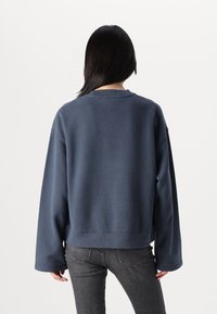 Sweat-shirt bleu marine en tissu doux, avec une coupe oversize, des manches larges et un col côtelé. Porté avec un jean gris foncé.