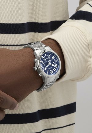 Srebrna ura iz nerjavečega jekla z velikim temno modrim številčnicom, belimi številkami in tremi podštevilčnicami. Nosi se na zapestju s črtastim puloverjem.