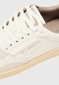 White sneaker with light beige accents, featuring laces, a textured suede toe cap, and a logo in green on the side. Rubber sole.