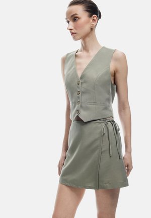 Completo gilet e gonna verde oliva realizzato in tessuto morbido. Il gilet presenta una profonda scollatura a V e bottoni, mentre la gonna ha una cintura regolabile.