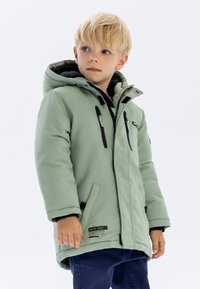 Veste matelassée vert clair avec des accents noirs, capuche et fermeture éclair sur le devant. Comprend deux poches latérales et une étiquette logo à l'ourlet.