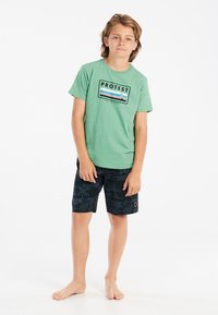 Groene katoenen T-shirt met "PROTEST" grafiek; zwarte zwembroek met palmbomendessin; blote voeten. Eenvoudige, casual strandoutfit.