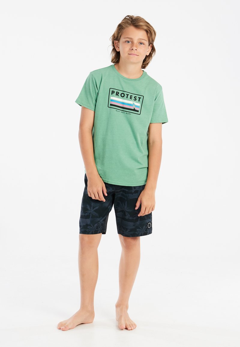 Groene katoenen T-shirt met "PROTEST" grafiek; zwarte zwembroek met palmbomendessin; blote voeten. Eenvoudige, casual strandoutfit.