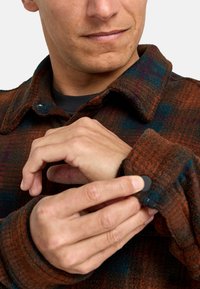 Karierte Flanellhemd in Braun, Orange und Blau. Verfügt über einen Kragen, eine Knopfleiste und einen strukturierten Stoff. Hände passen den Ärmel an.