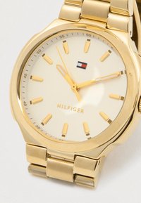Kuldtooni Tommy Hilfigeri randmevaat, millel on kreemivärvi kelladial, kuldse numbrite ja kätega ning ühendatud metallist käevõru, mis näitab aega 10:14:57.