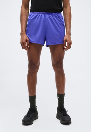 Mees kannab lilla sportlühikeseid pükse, musti tossusid, tumerohelisi sokke ja musta tuba, seisab tavalise heleda tausta ees.