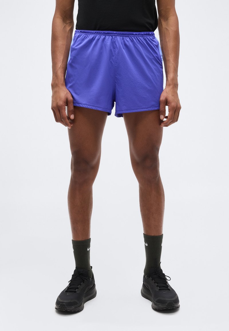 Άνδρας φοράει μωβ αθλητικές σορτς, μαύρα αθλητικά παπούτσια, σκουροπράσινες κάλτσες και μαύρο μπλουζάκι, στέκεται μπροστά σε απλό φωτεινό φόντο.