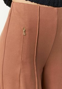 Pantalons taille haute en couleur rouille, dotés d'une fermeture éclair dorée et d'un tissu lisse et extensible avec des coutures verticales pour un look ajusté.