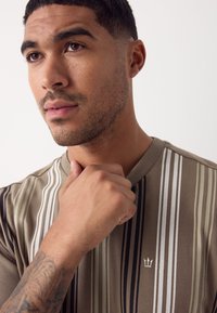 Csíkos rövid ujjú ing barna, bézs és fehér földszínekkel. Kerek nyakkivágással és hímzett logóval a mellkason.