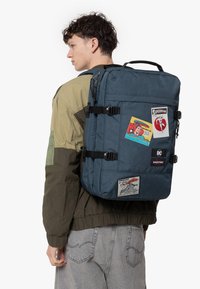 Blue, textured fabric backpack with multiple patches and tags. Features adjustable straps, a top handle, and a rectangular shape.