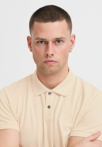 Kortermet beige polo-skjorte med strukturelt stoff, to mørke knapper på kragen og en diskret logo på brystet.