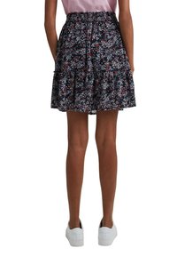 Femme portant une mini-jupe noire à imprimé floral avec un ourlet à volants, un top rose et des baskets blanches, debout, dos à la caméra.