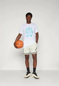 T-shirt branco da Nike com gráfico aqua, calções com padrão preto e branco, segurando uma bola de basquete, a usar meias pretas e ténis creme.