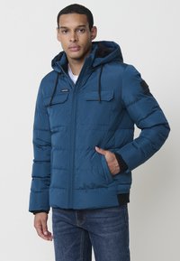 Chaqueta acolchada azul con capucha. Presenta dos bolsillos en el pecho, puños elásticos y cierre de cremallera. El material parece suave y resistente al agua.