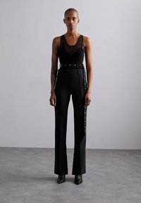Black sleeveless top with a sheer overlay paired with high-waisted black trousers featuring side snaps and a wide belt, finished with pointed heels.
