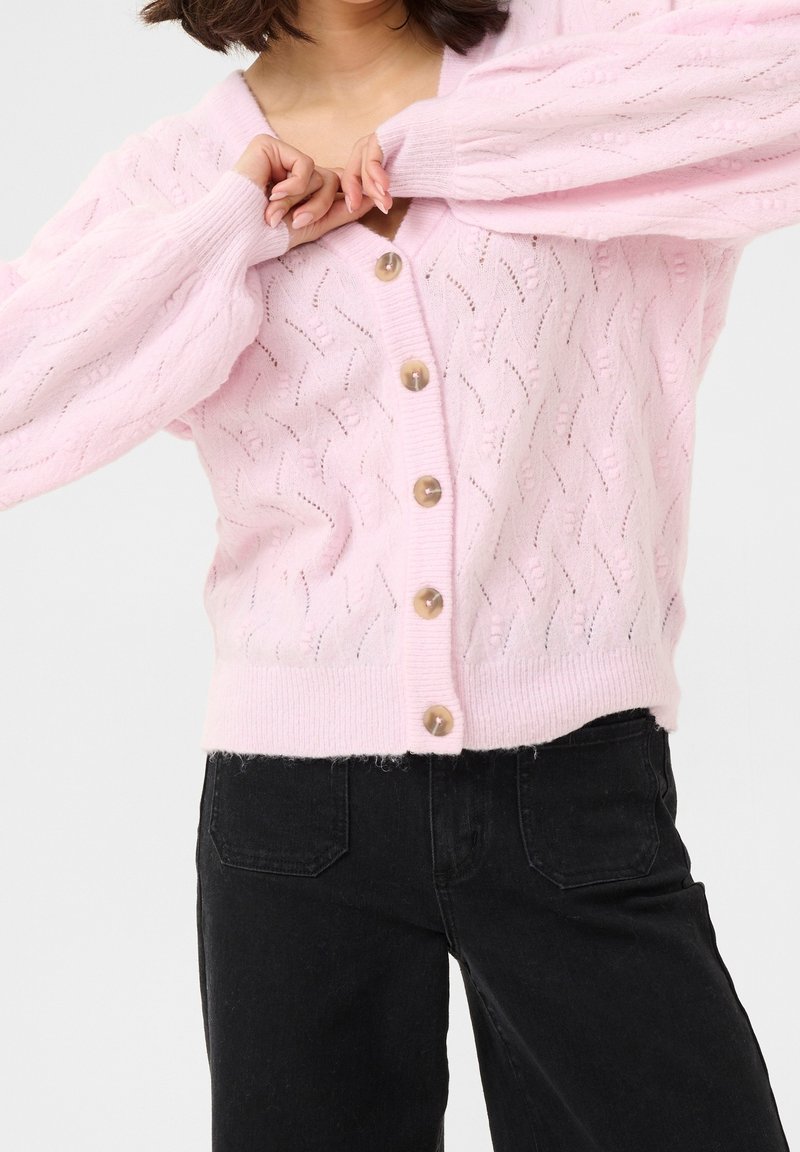 Femme portant un cardigan tricoté rose clair avec des boutons en bois et un pantalon noir, ajustant le cardigan près du col.