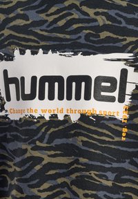 Czarna koszulka z oliwkowozielonym wzorem w tygrysie paski; posiada wyraźne logo "hummel" oraz pomarańczowy tekst "Zmień świat poprzez sport."