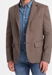 Brązowa marynarka w kratę z klapami z wycięciem, pojedynczą kieszenią na piersi i dwoma przednimi kieszeniami, nałożona na niebieską koszulę dżinsową. Gładka faktura tkaniny.