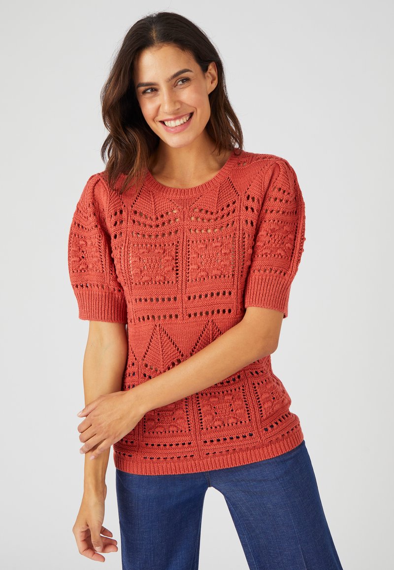 Maglione lavorato a maglia in corallo con maniche a sbuffo corte, motivi geometrici testurizzati e dettagli ad intaglio. Abbinato a pantaloni di jeans blu.