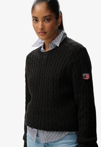 Zwarte kabelgebreide trui met een ronde halslijn en een logo-patch met een Amerikaanse vlag op de linkermouw. Draag het over een gestreepte shirt.